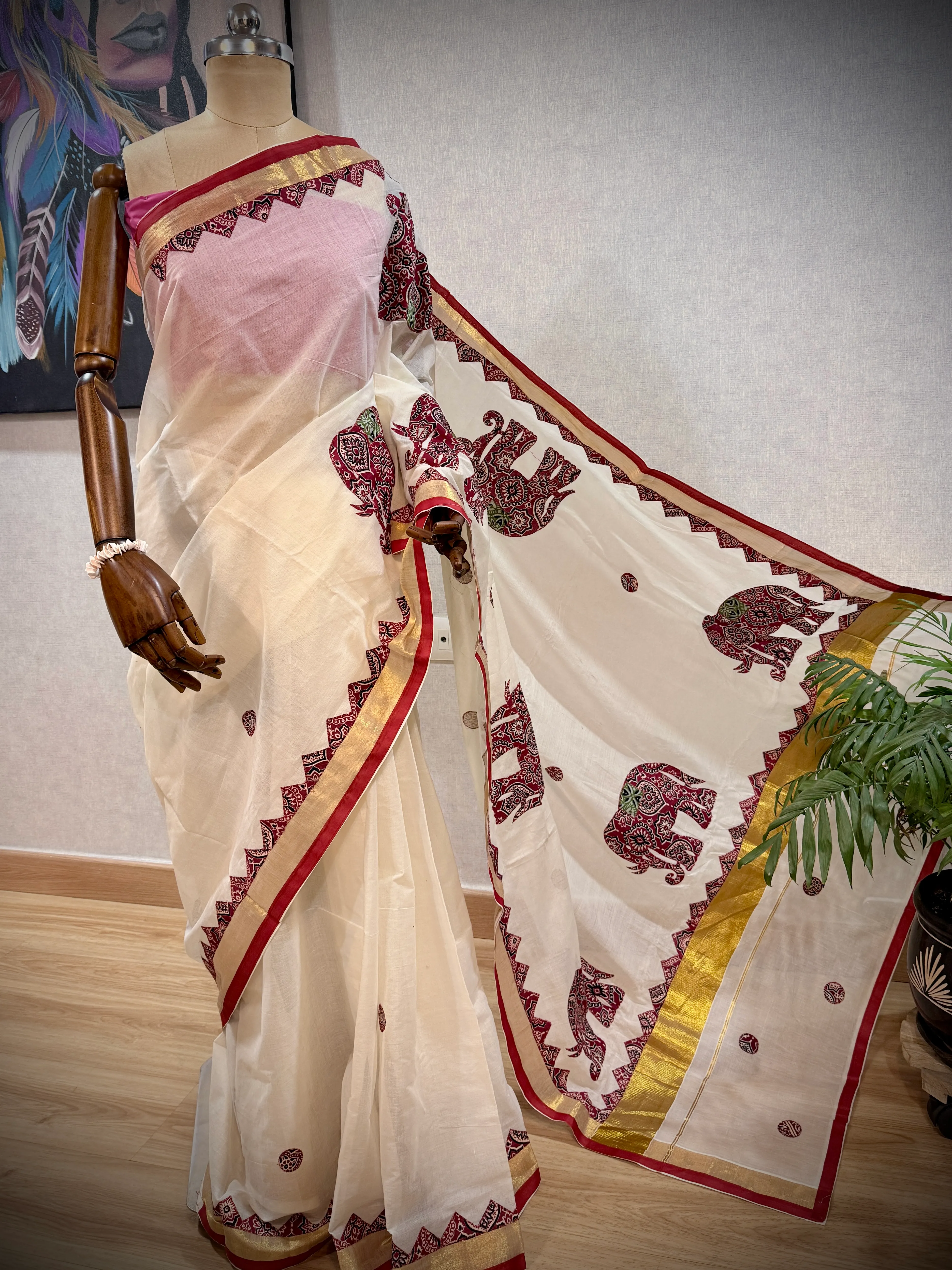 Cream Kerala cotton saree with maroon appliqué elephant motifs on the pallu, draped on a mannequin, close vertical crop showing the shoulder drape and gold-edged border.