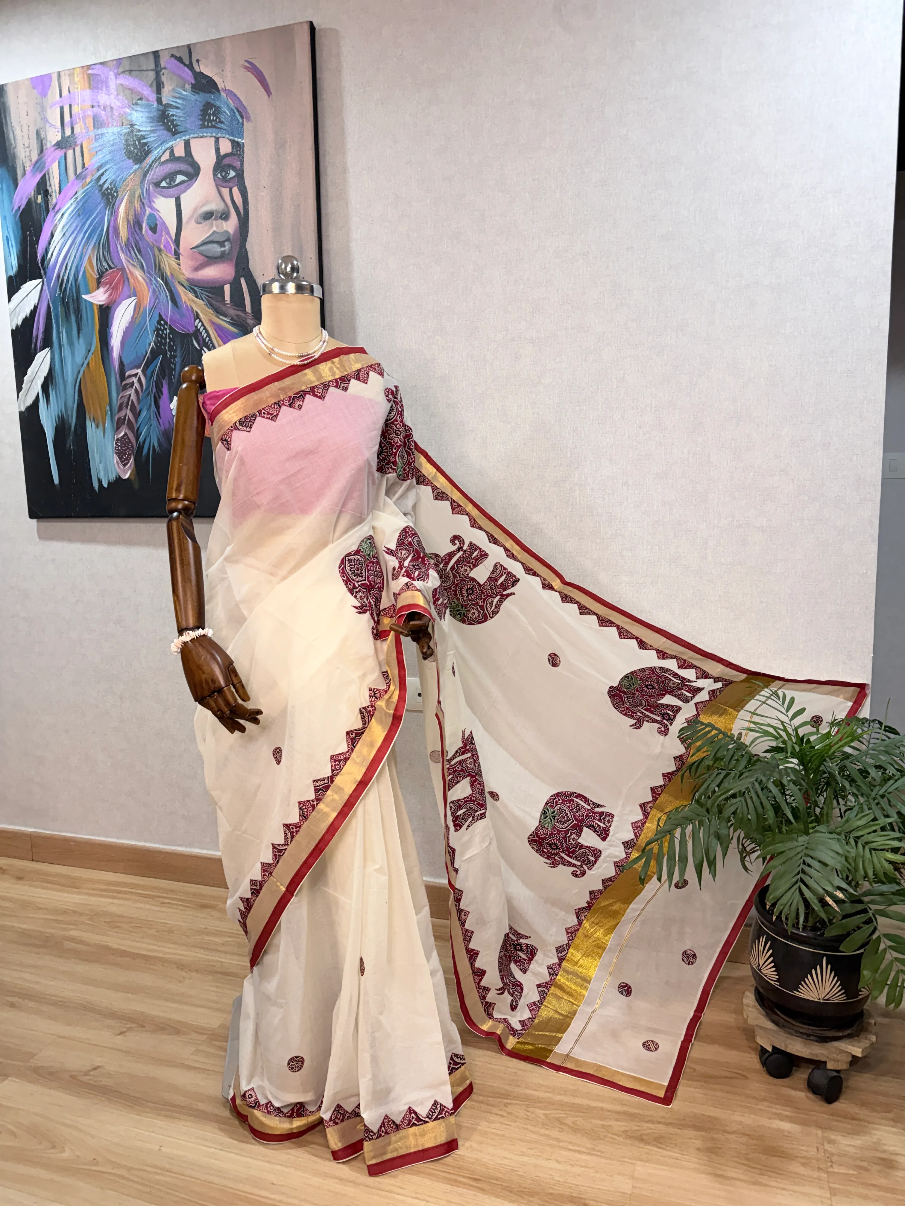 Cream Kerala cotton saree on the mannequin from a front three-quarter angle, pallu fanned open beside a potted palm and a feather-motif painting in the background.