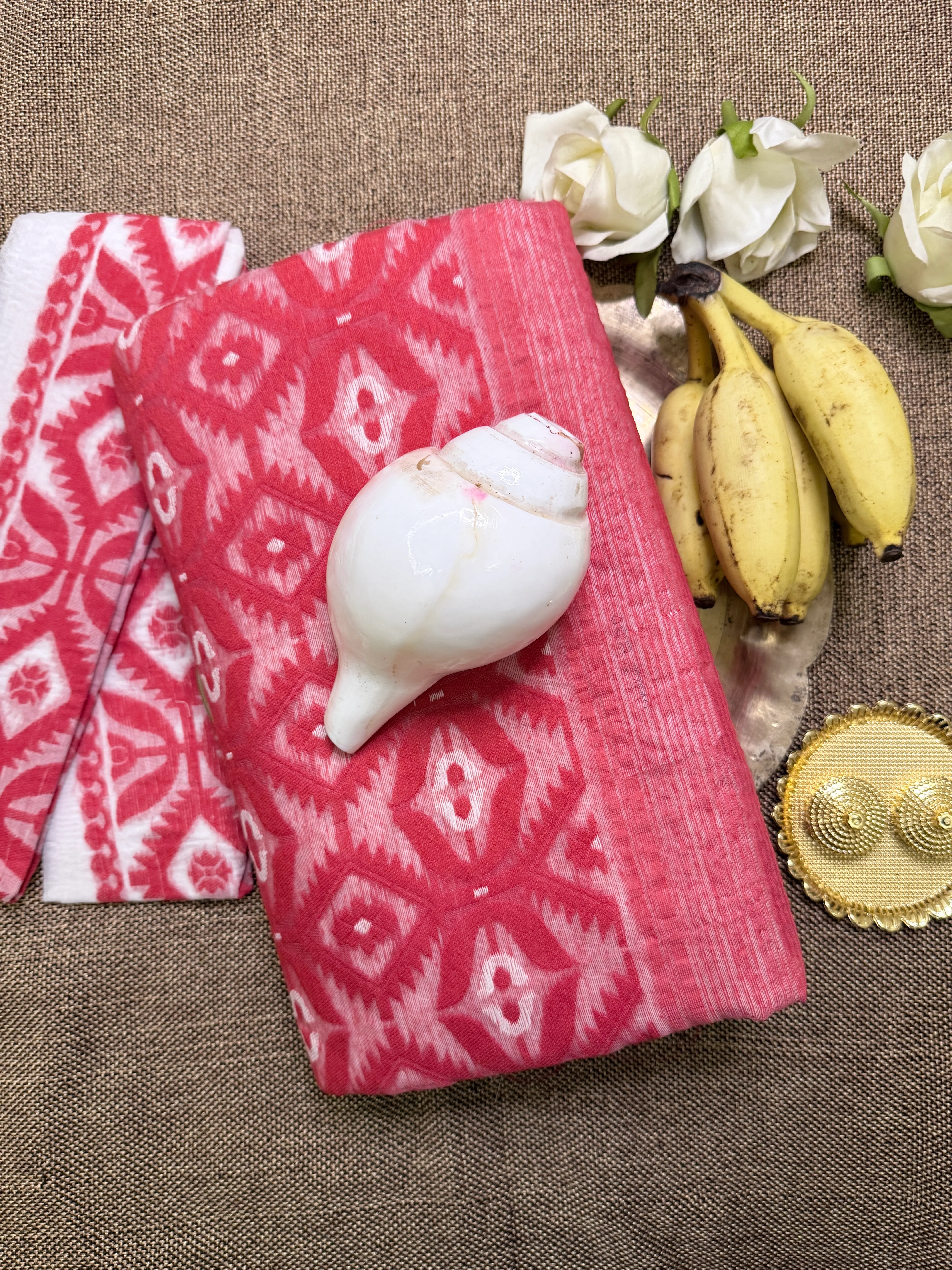 Two folded pink-and-white Dhakai jamdani sarees with an ikat-style diamond motif, styled with bananas, white roses and a conch shell for a puja flat-lay.