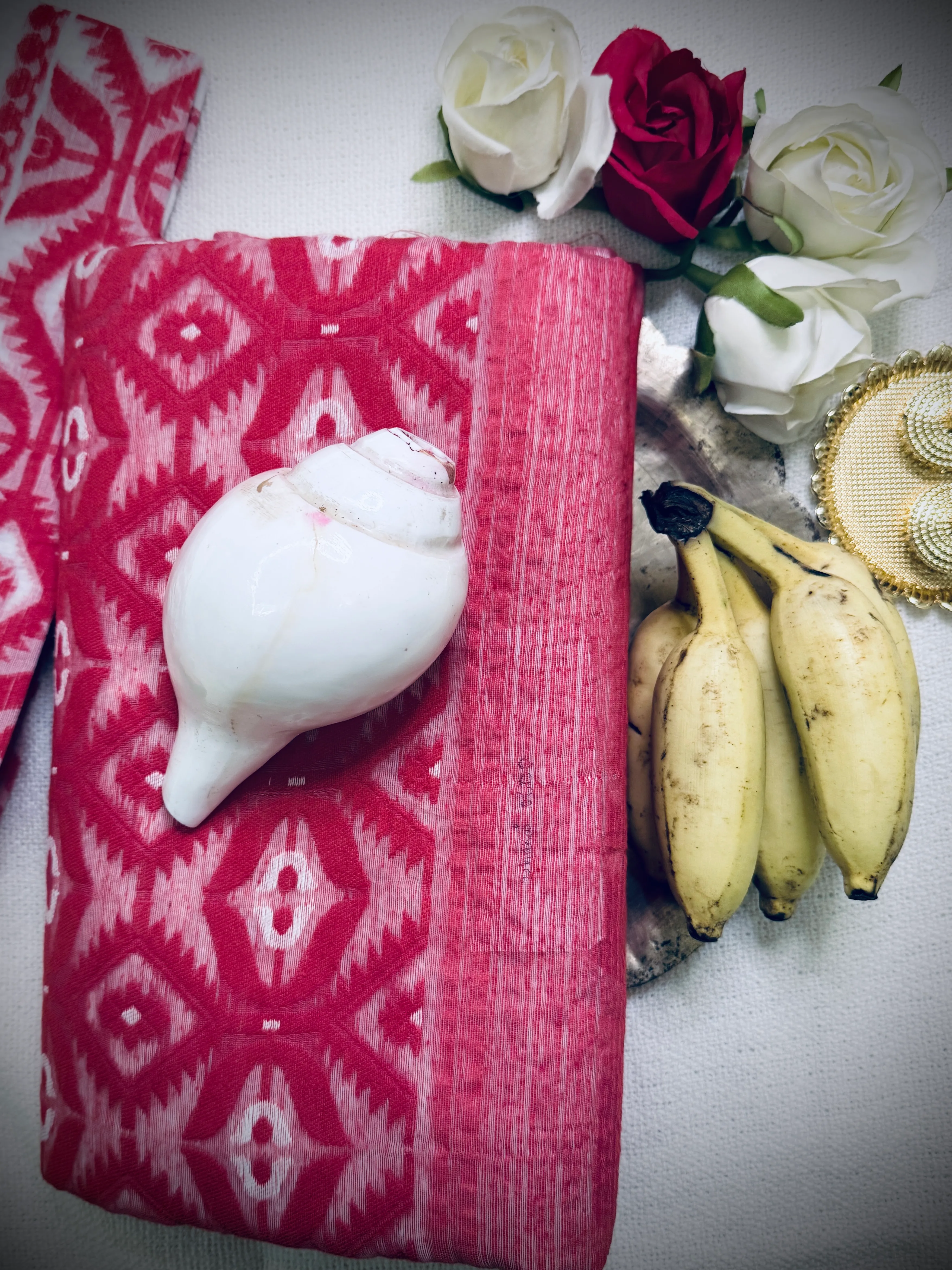 Folded pink-and-white Dhakai jamdani saree with ikat diamond motifs, close flat-lay with white and red roses, bananas and a conch shell.
