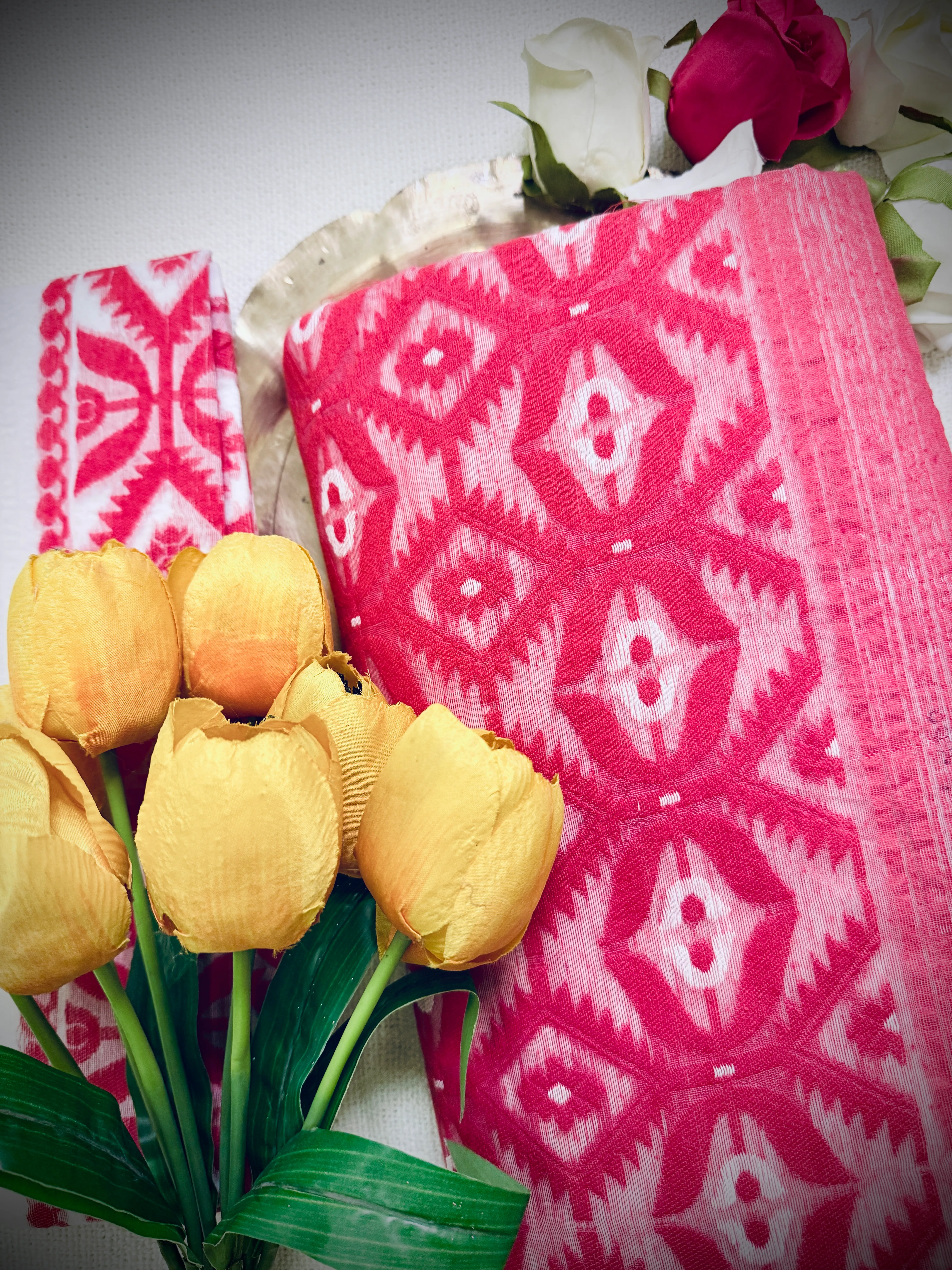 Folded Dhakai jamdani saree showing the red ikat diamond pattern against white, styled with yellow tulips.