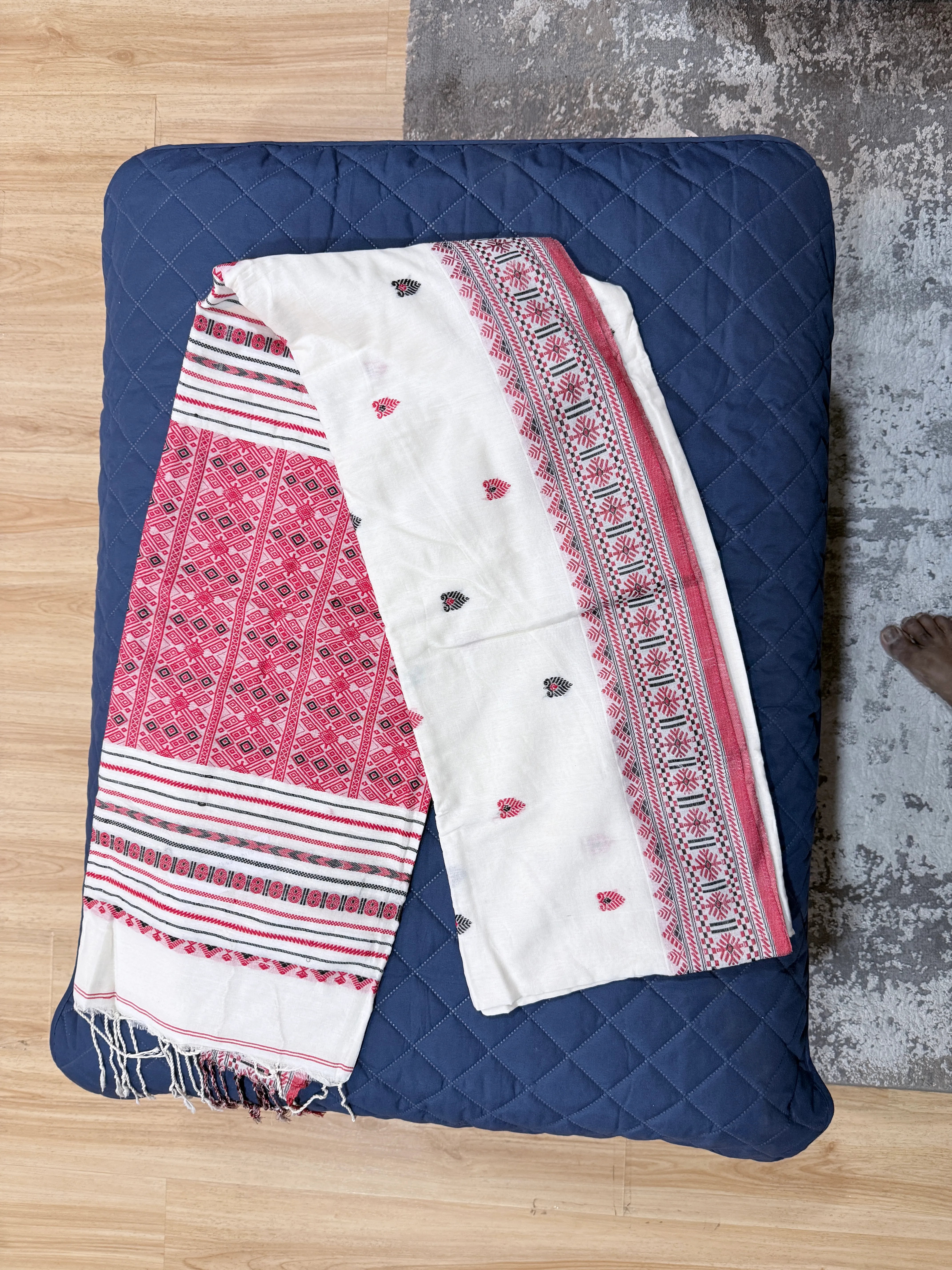 Folded Dhonekhali cotton saree with a pink-and-black woven geometric border and cream body, laid flat on a navy quilted surface.