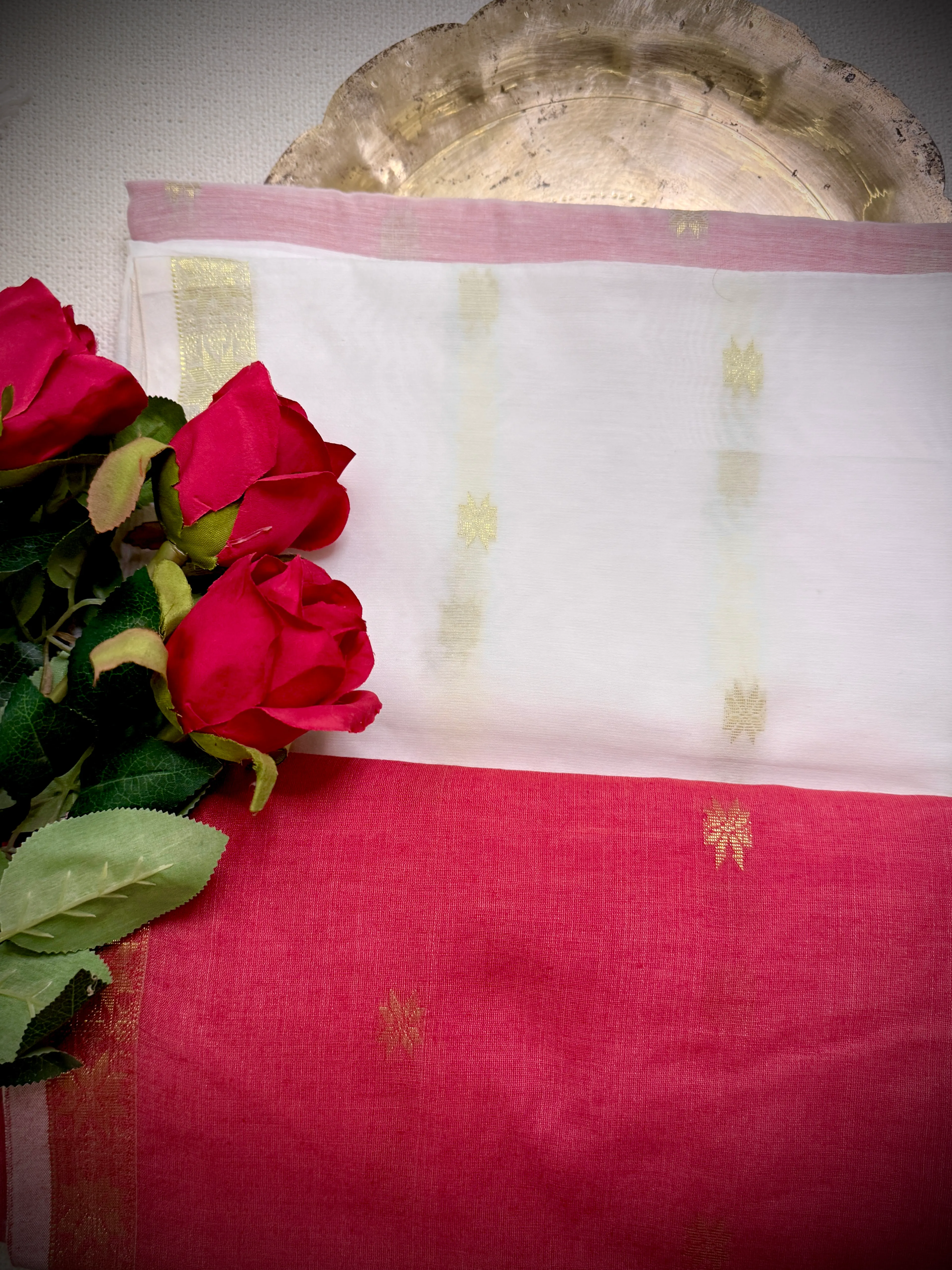 Folded white-and-pink khadi cotton saree with scattered gold star motifs and a plain pink-red body, styled with red roses beside a gold plate.