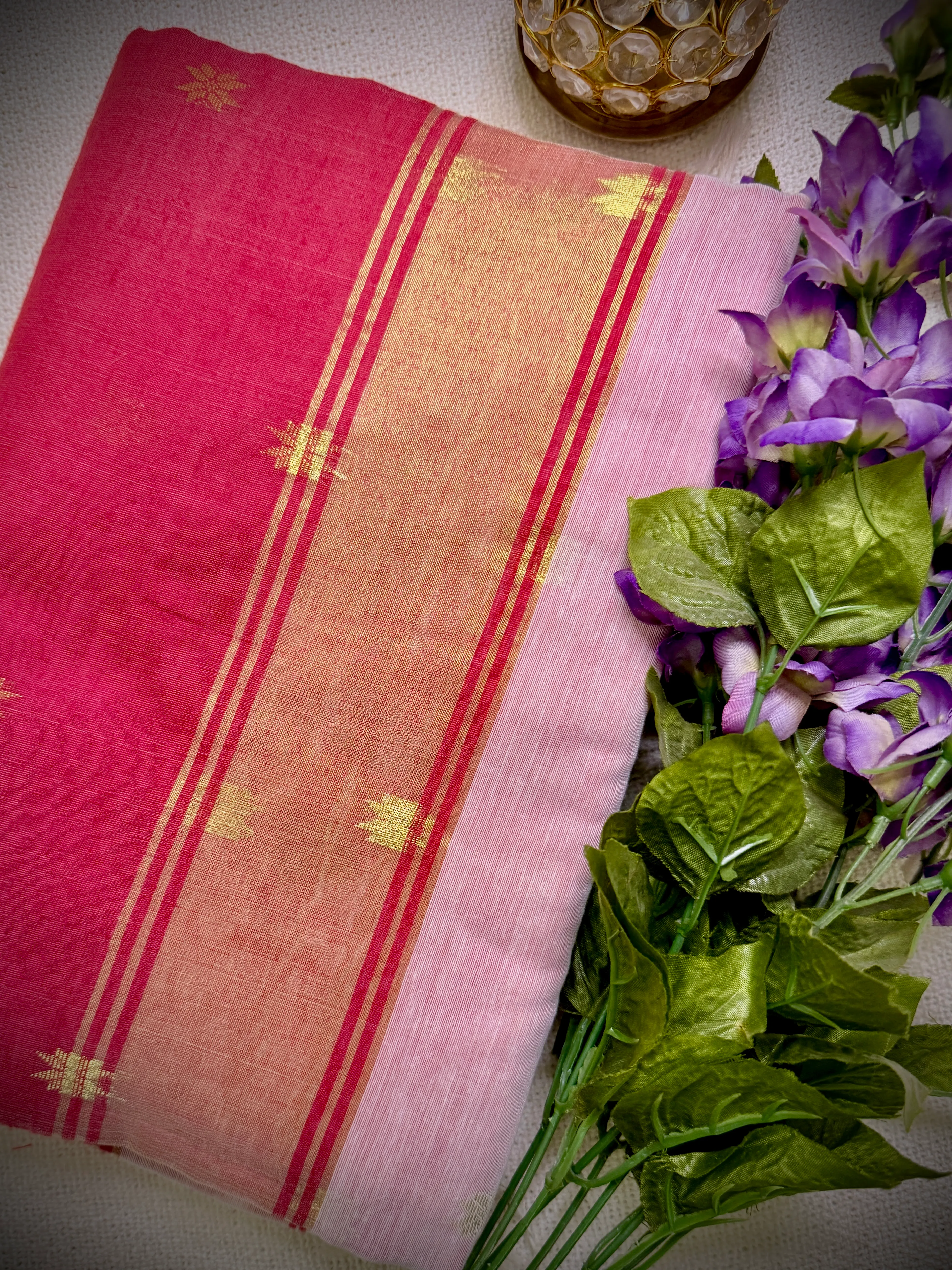 Khadi cotton saree's pink-red pallu with a gold zari border, styled with purple flowers and a gold candle holder.
