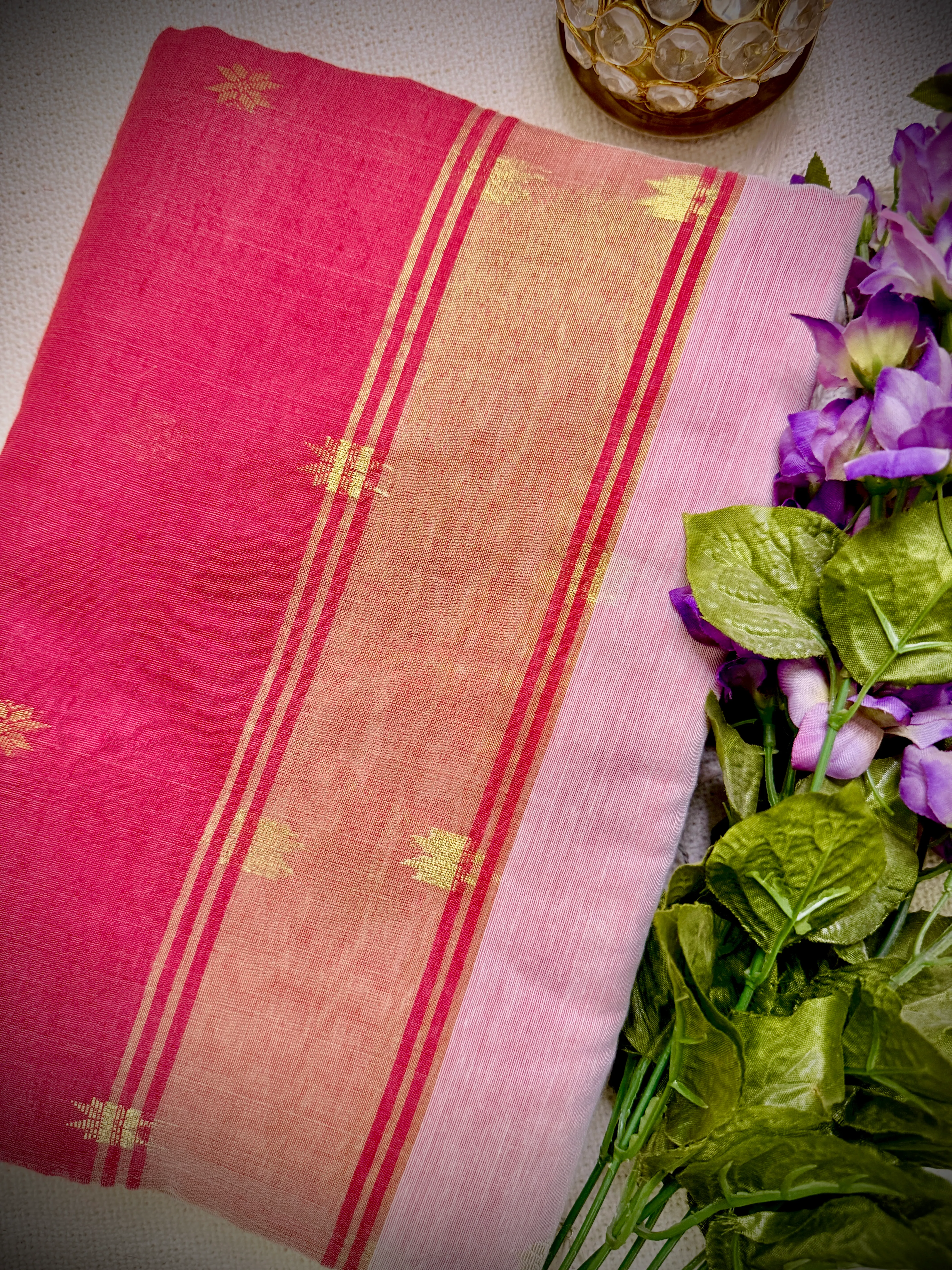 Khadi cotton saree's pink-red pallu with gold zari border and star motifs, styled with purple flowers and a gold candle holder.