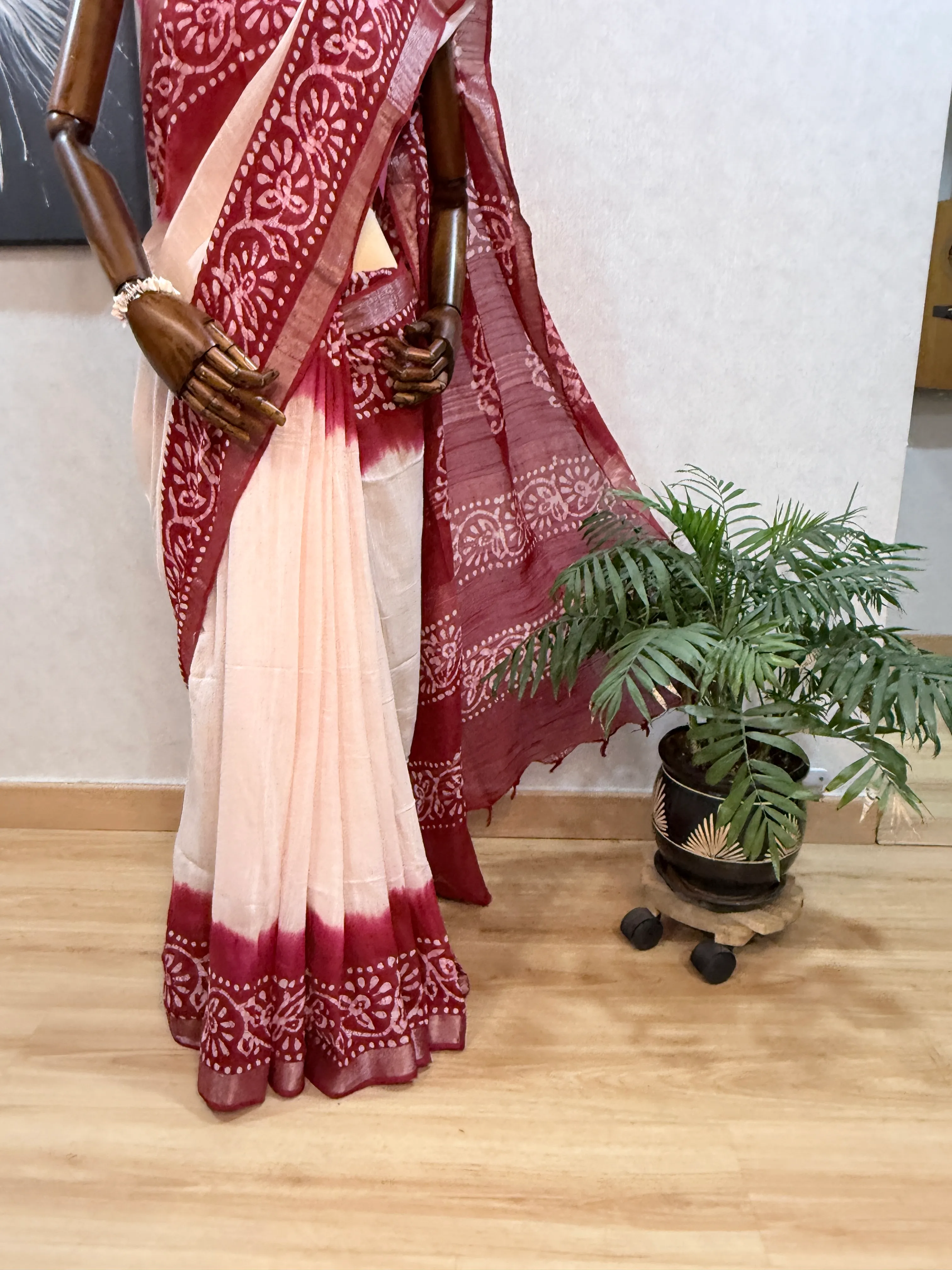 Maroon batik-print linen saree on the mannequin, closer framing of the pallu and pleats beside the same palm.