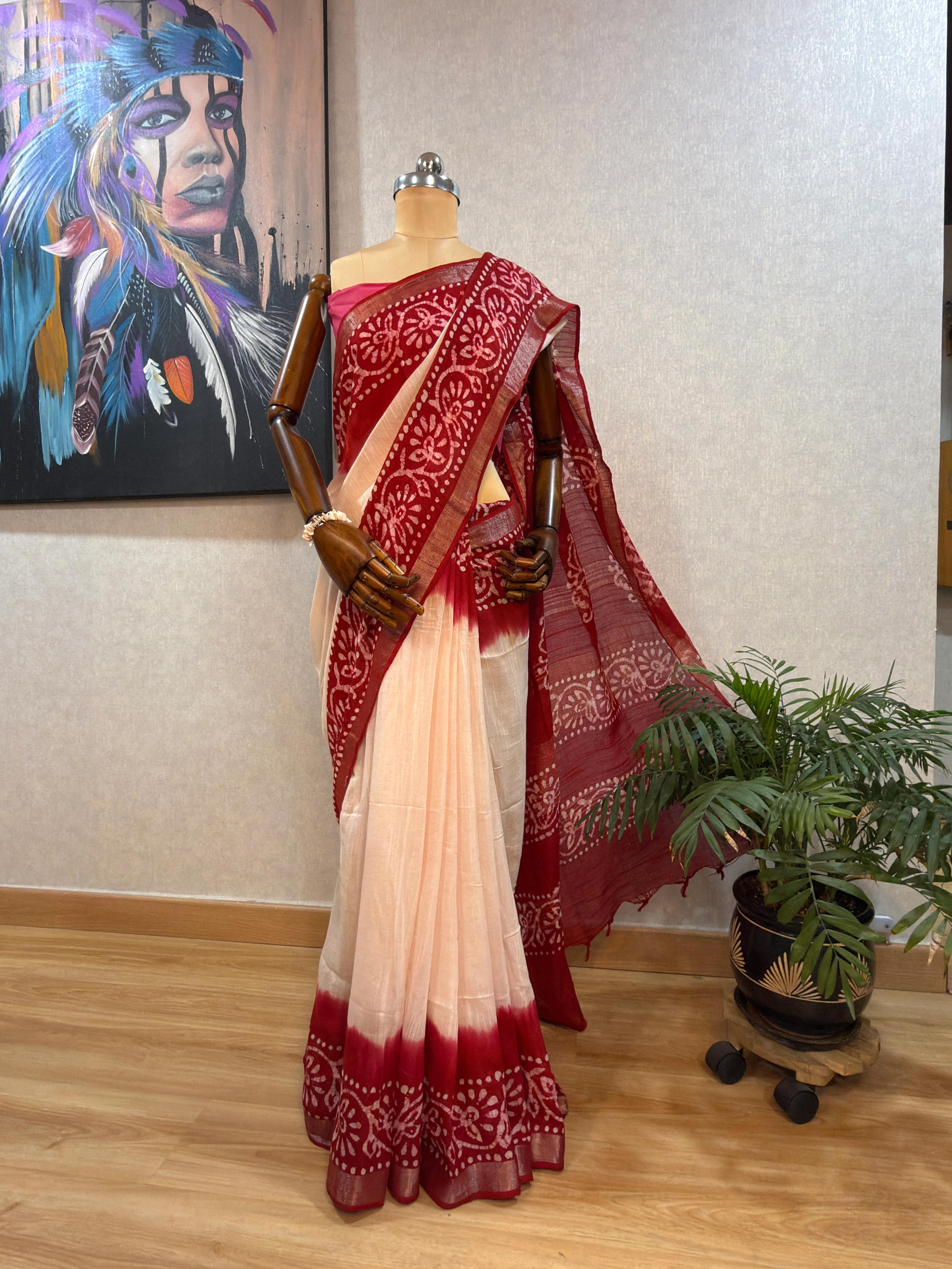 Maroon batik-print linen saree on the mannequin, full-length view beside a potted palm and a feather-motif painting.