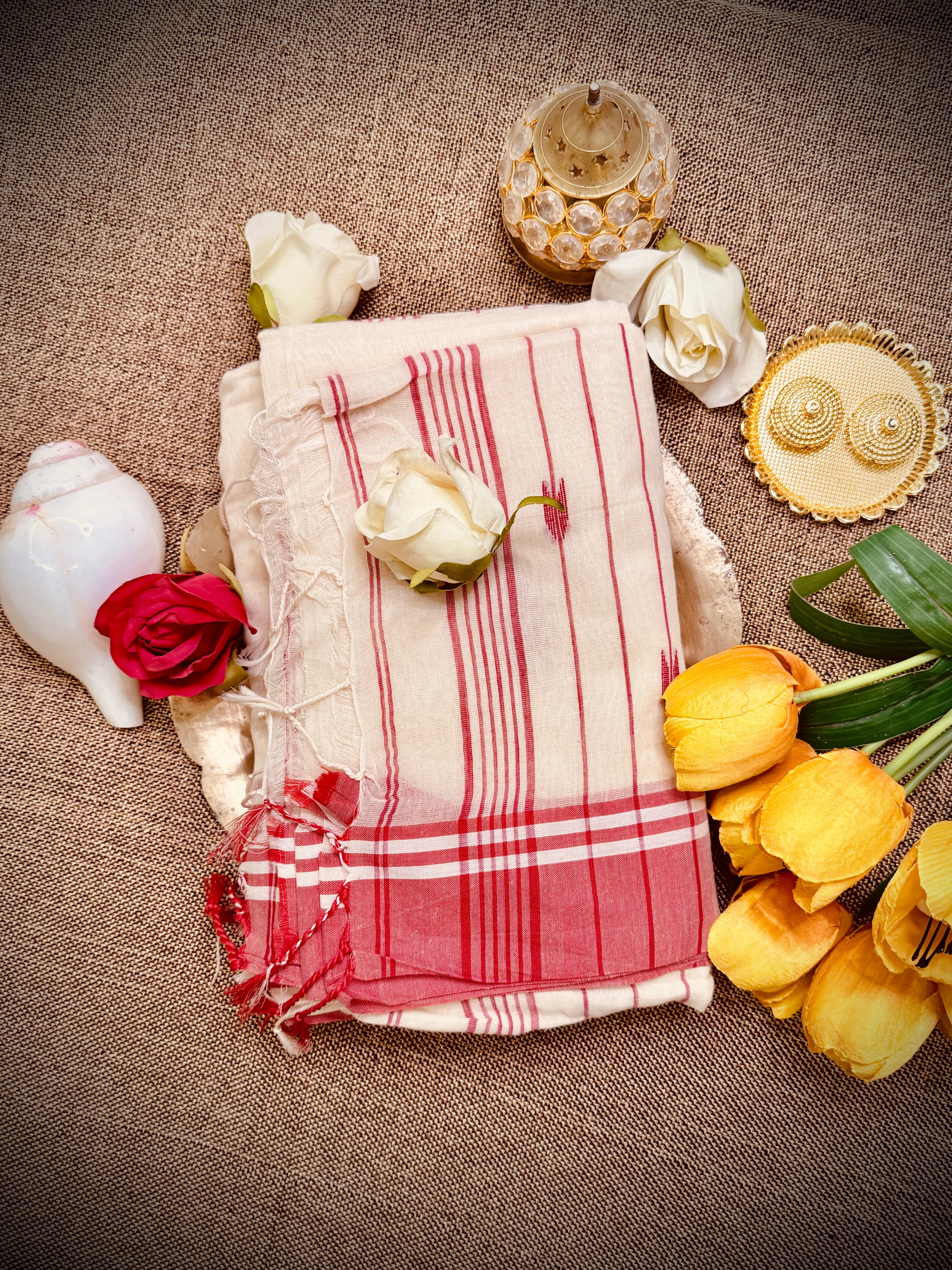 Folded cream mul cotton Tangail saree with red stripes and plaid border, styled with roses, a conch shell and a brass lamp.