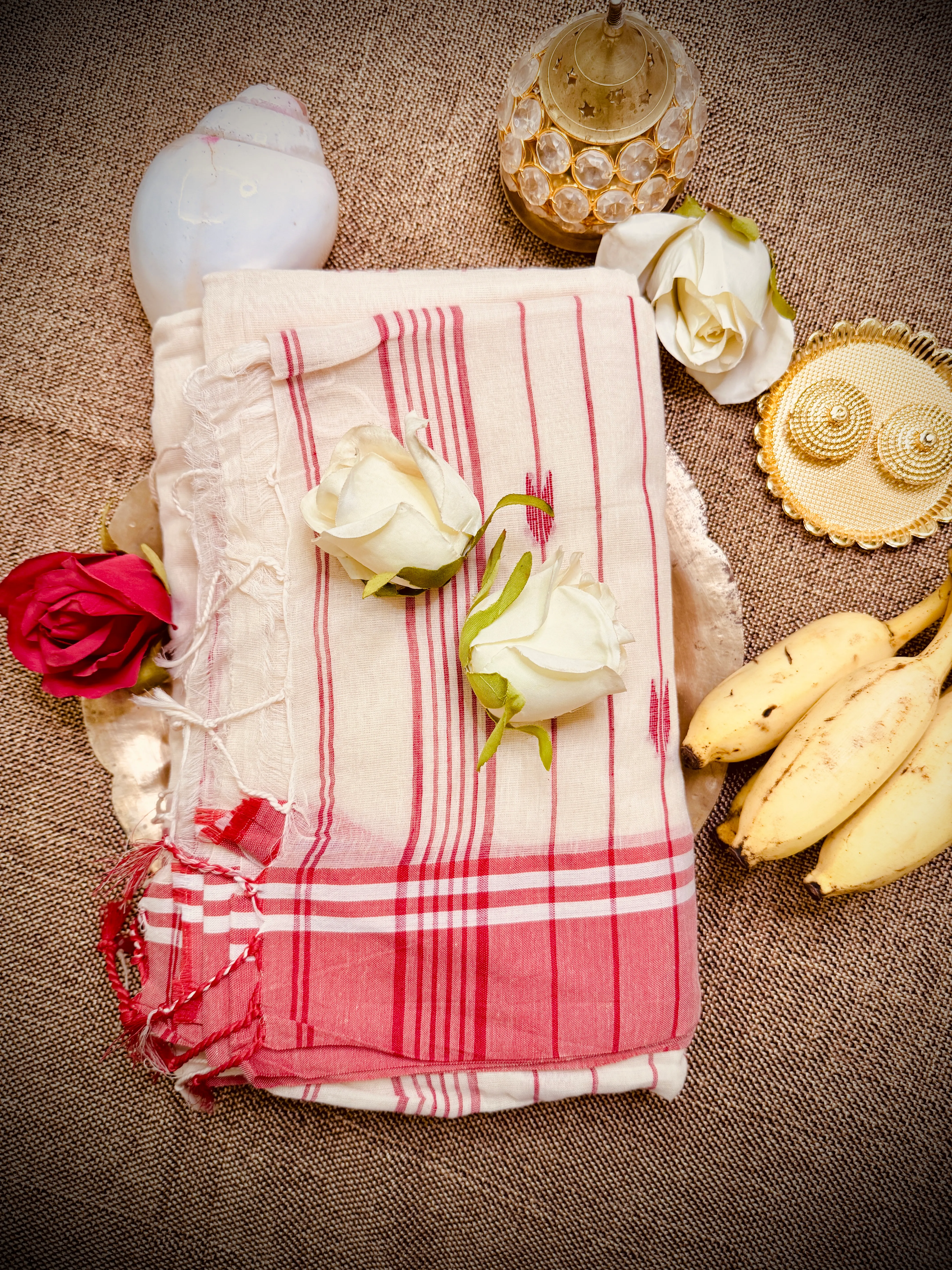 Folded cream mul cotton Tangail saree with red stripes and plaid border, styled with white roses, bananas and a brass lamp.