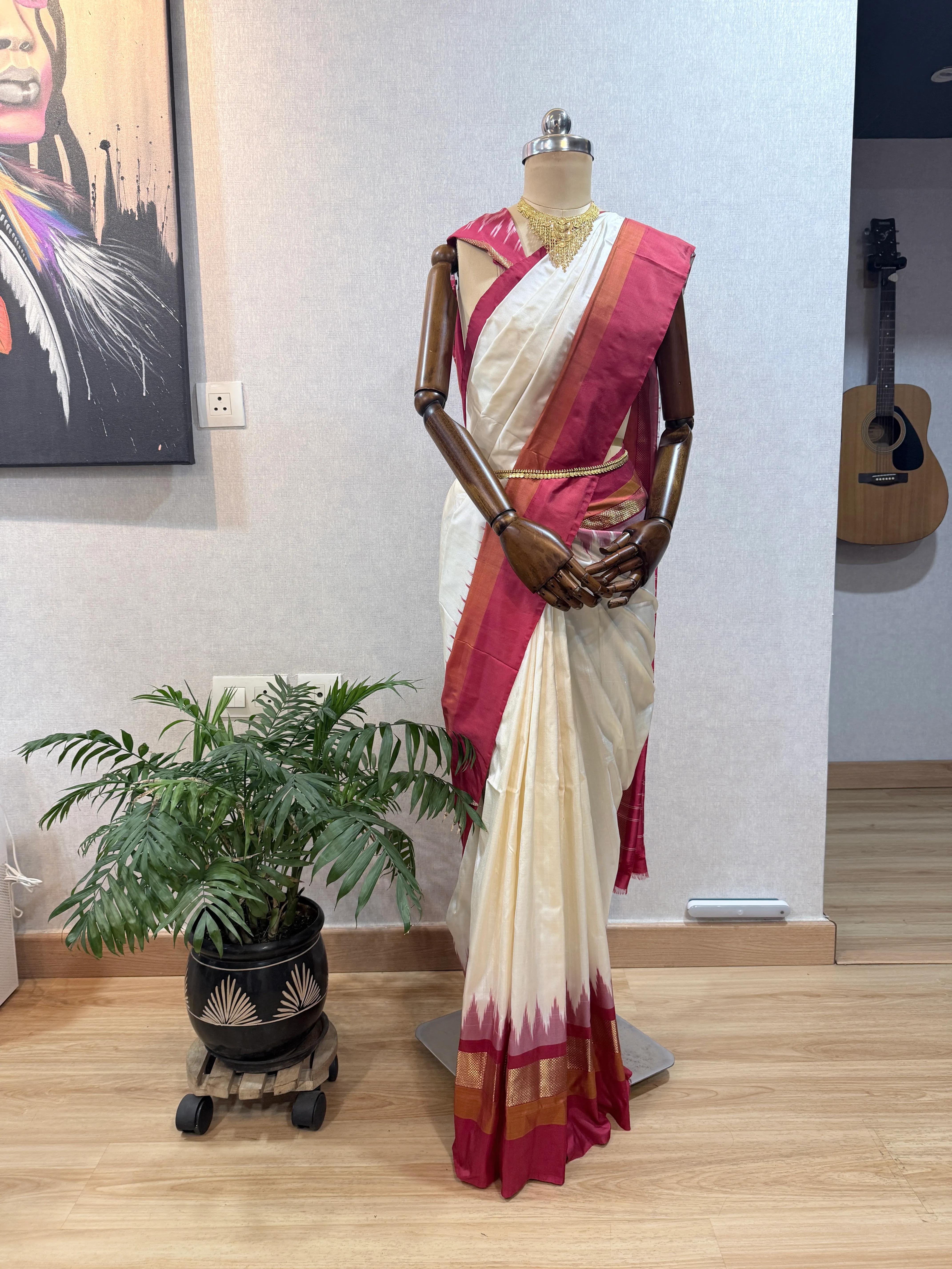 Ivory saree with a maroon-and-gold ikat border draped on a mannequin, full-length view beside a potted palm.